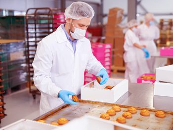 Confectionery factory worker in white coat collecting freshly baked pastry from tray and putting it into paper box.