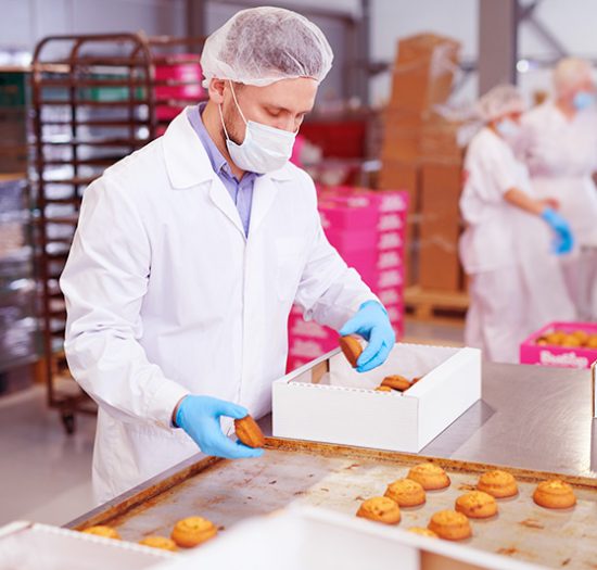 Confectionery factory worker in white coat collecting freshly baked pastry from tray and putting it into paper box.